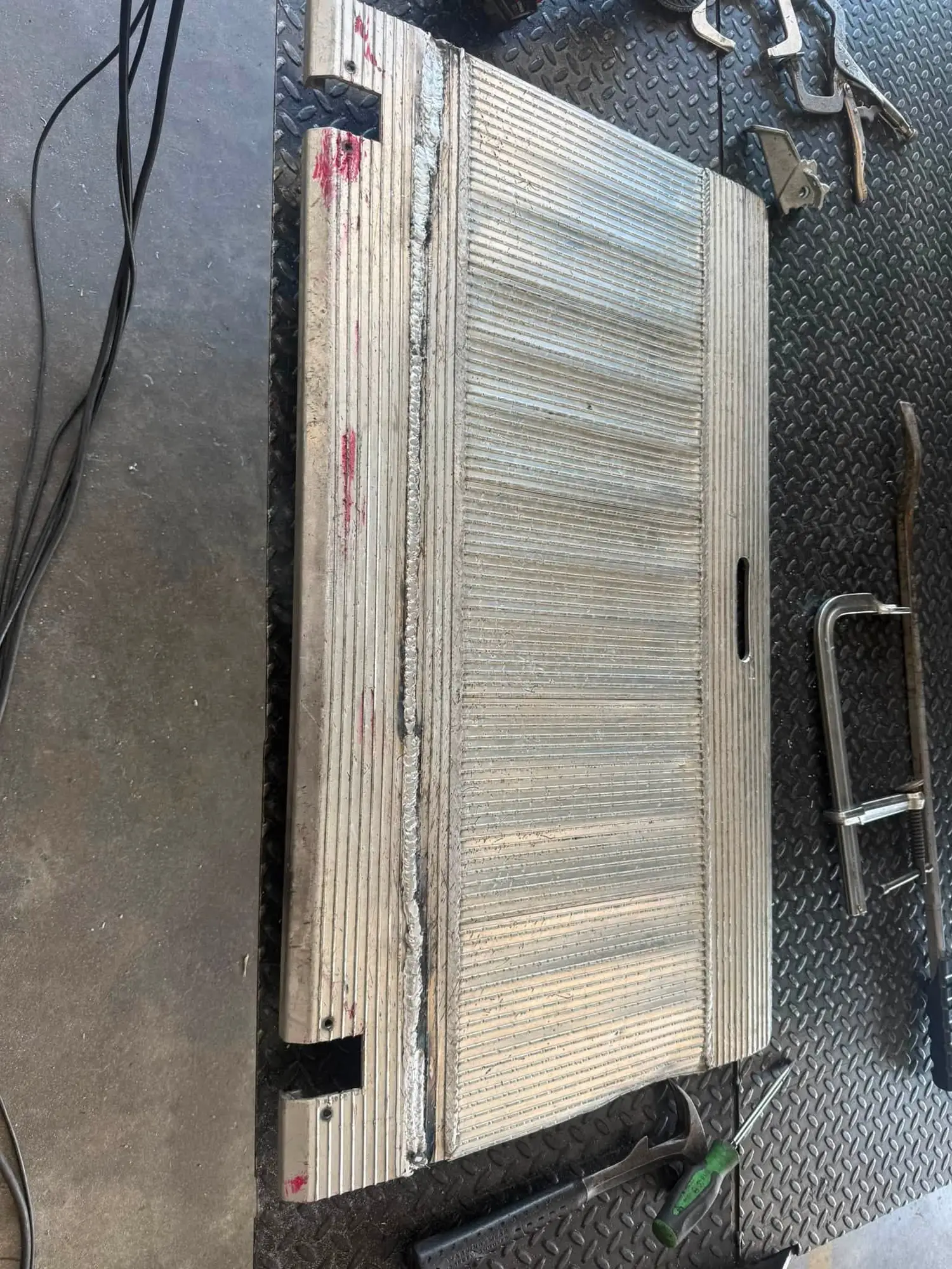 A liftgate platform section laid on a diamond plate work surface with a fresh weld seam and C-clamps beside it